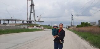 A man stands holding a child in front of a bridge that is not completed.