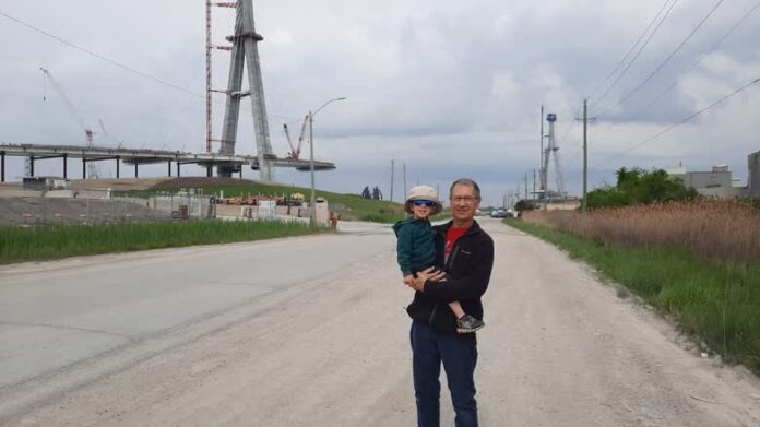 A man stands holding a child in front of a bridge that is not completed.