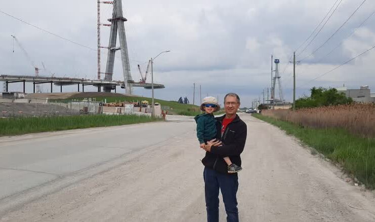 A man stands holding a child in front of a bridge that is not completed.