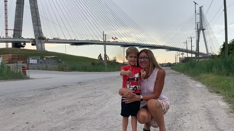 A child stands with his thumbs and a woman is crouching beside him, with the bridge in the background.
