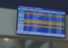 A screen hanging on a wall shows a list of international arrivals from destinations like Amsterdam, Barcelona and London.