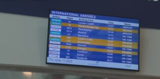 A screen hanging on a wall shows a list of international arrivals from destinations like Amsterdam, Barcelona and London.