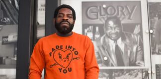 A headshot of man wearing an orange shirt against a black and white background.