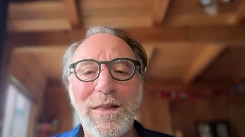 A man wearing a blue shirt and glasses in a wooden cabin.