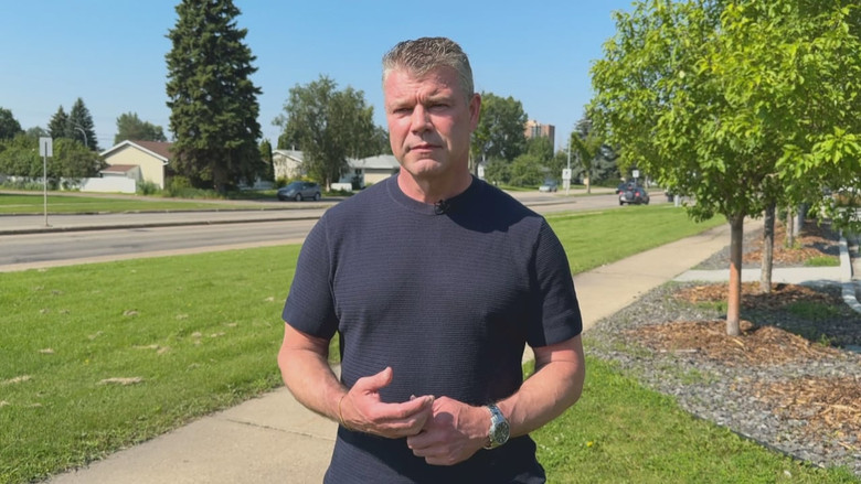 A white man stands in front of a camera. He wears a dark blue shirt and a watch. Behind him is a residential neighbourhood.