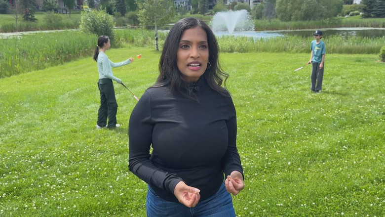 A woman with long black hair stands in front of a green space while children play behind her. She is wearing jeans and a black turtleneck.