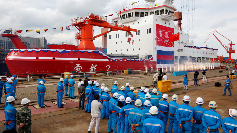 Many people in blue overalls and white helmets stand on a dock in front  of a large red and white ship.