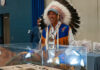 A man wearing an Indigenous headdress, white shirt and blue and brown leather vest smiles as he removes a blanket covering a display case with artifacts.