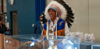 A man wearing an Indigenous headdress, white shirt and blue and brown leather vest smiles as he removes a blanket covering a display case with artifacts.