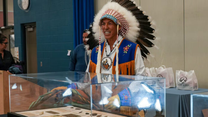 A man wearing an Indigenous headdress, white shirt and blue and brown leather vest smiles as he removes a blanket covering a display case with artifacts.