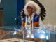A man wearing an Indigenous headdress, white shirt and blue and brown leather vest smiles as he removes a blanket covering a display case with artifacts.