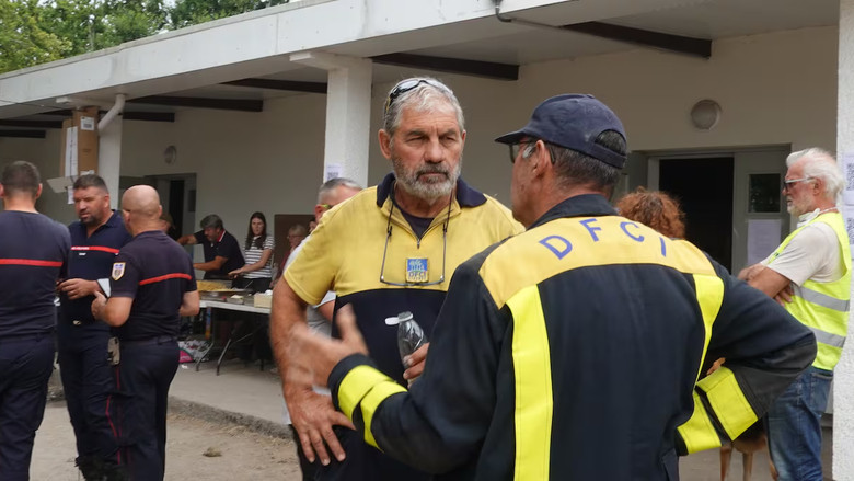 A man with grey hair and a yellow polo holds his hands on his hips and speaks to another man.