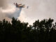 A yellow plane dumps water over a forest in a smoky sky.