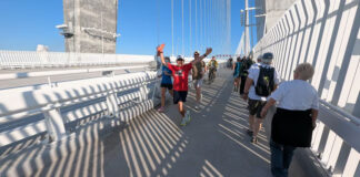 A man in a t-shirt with his arms in the air in a welcoming stance leads a row of people walking down the path while another row walks in the other direction.