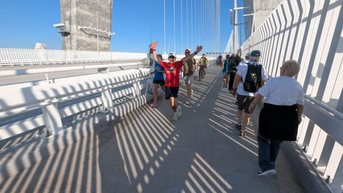 bridgef-001 A man in a t-shirt with his arms in the air in a welcoming stance leads a row of people walking down the path while another row walks in the other direction.