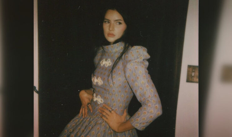A young woman with dark hair and a long dress poses for the camera in a promotional photo.
