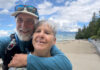 A smiling couple embraces while taking a selfie beside the seawall and beach in Vancouver’s Stanley Park.