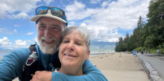 A smiling couple embraces while taking a selfie beside the seawall and beach in Vancouver’s Stanley Park.