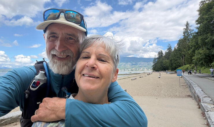 A smiling couple embraces while taking a selfie beside the seawall and beach in Vancouver’s Stanley Park.