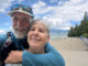 A smiling couple embraces while taking a selfie beside the seawall and beach in Vancouver’s Stanley Park.