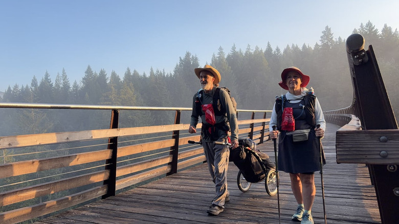 Two hikers wearing backpacks and sun hats walk across a trestle bridge with trekking poles.