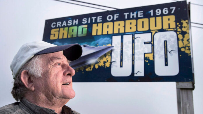 A man is shown in front of a billboard that says 'Crash site of the 1967 Shag Harbour UFO.'