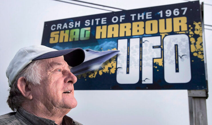 A man is shown in front of a billboard that says 'Crash site of the 1967 Shag Harbour UFO.'