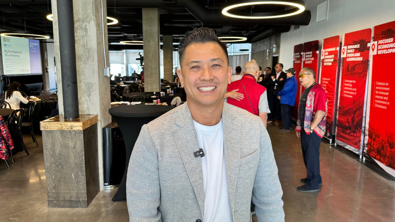 A smiling man looks at the camera and stands in a room with people behind him.