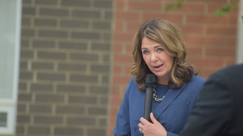 A woman speaks into a microphone in front of a dark, grey brick wall.